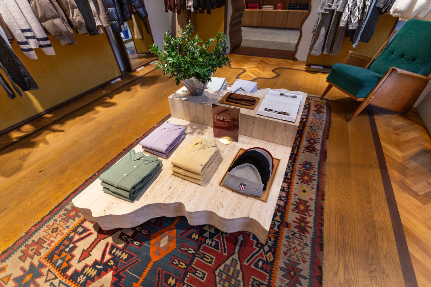 Gant Store'i näidissalongis laua peal on triiksärgid ja sallid, taustal puidust põrand ja rohekas-taim, värvikirev vaip ning ruumi pehme valgustus, Gant ja Marmi Futerno viidetega details
