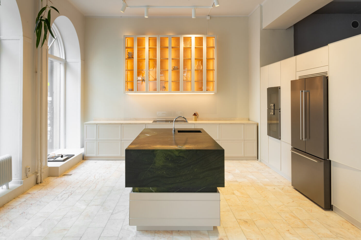 Lucks Showroom köög I: valged köögikapid ja keskne roheline marmorm saar, roostevabast terasest seadmed, suur külmkapp ning lumineeritud valgustus, sobiv kaasaegse interjööri näide.