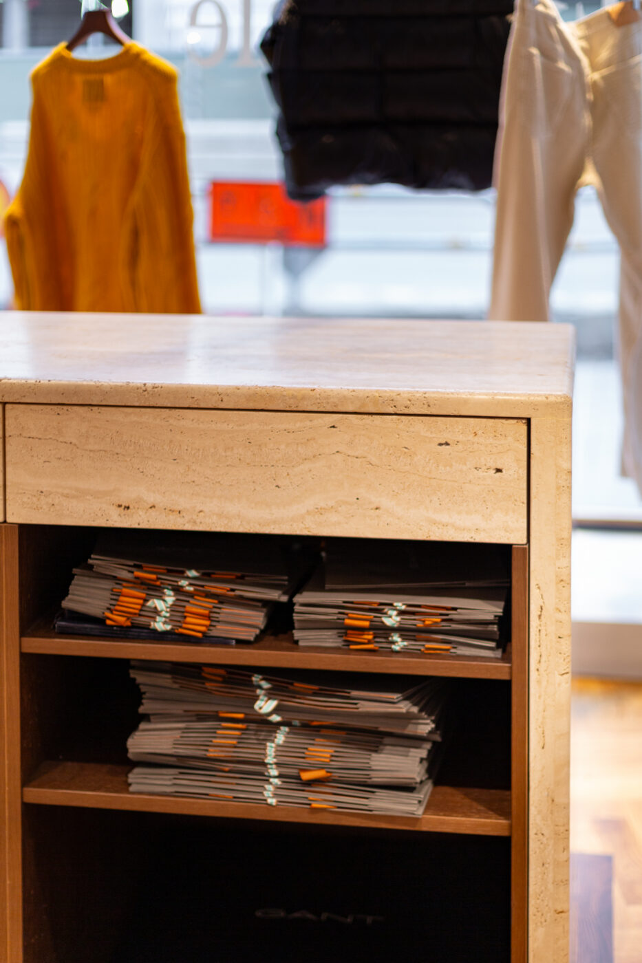 Kollane kleit ja musta ning valge pesuervisoonide taustal esinev puitlik kummut koos kahaneva raamatute ning ajakirjade virnadega; Gant Store stiiliga seotud kontekst.