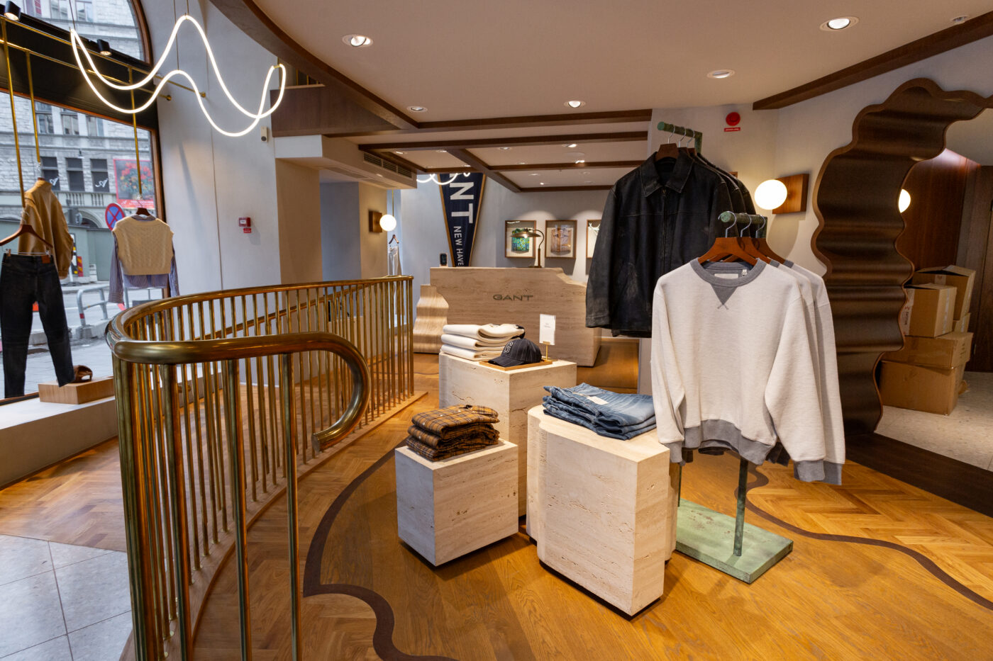 GANT Store Stockholmi interjöör: puidust põrand, lillelised ja minimalistlikud vitriinid, kerged riidepuud ja kollektsioonid nagu jakid ja Sweatshirtid; silma järgi eristuvad valguslambid ja kuldsed käsipliidid.