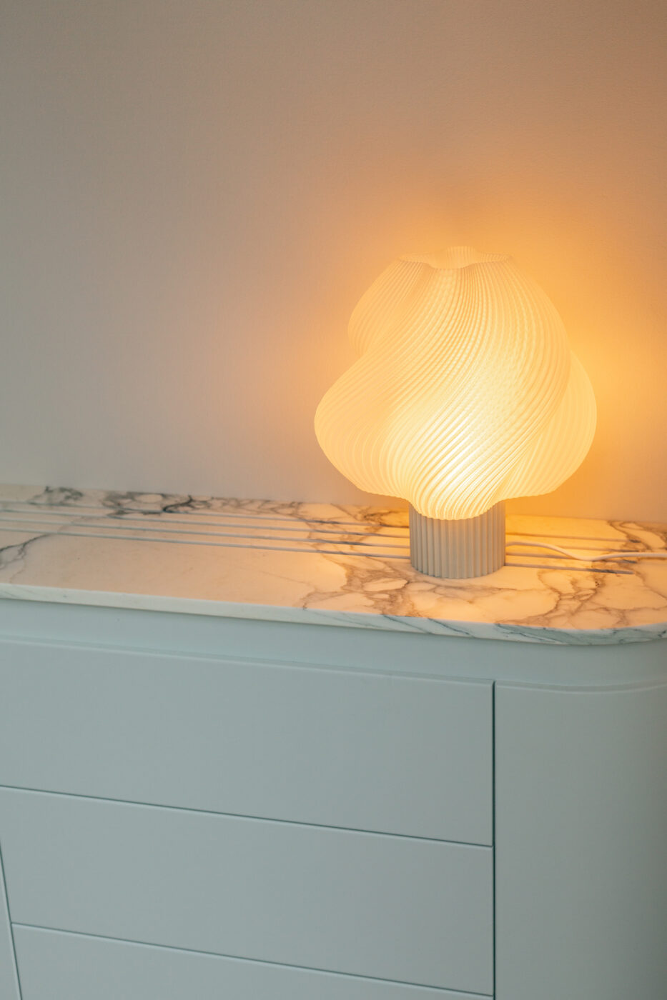 Valge marmormustriga kummutil asuv sooja valgusega kaasaegne lambihoone, sinine kapikarp ja hele taust, Apartment Callans projekti stiili visuaal. Marmi Futerno brändiga seostub lampli kaasaegne vorm.