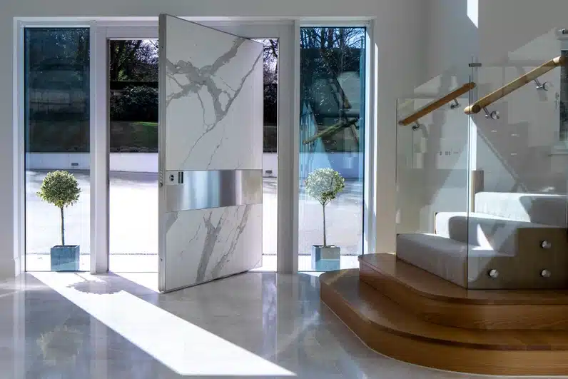 Statuario Altissimo marble pivot door in a modern home entryway with glass staircase and bright lighting.