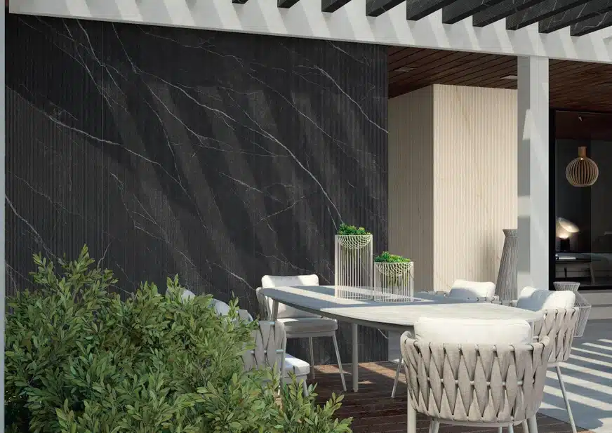 Calacatta Black Laminam musta marmorilaadse tekstuuriga pind, valge virvendus ja veenid Jälgib musta paneeli taustal mugulatud veranda stiilis välisruumi söögilaua ja valgete -toonidega toolid.