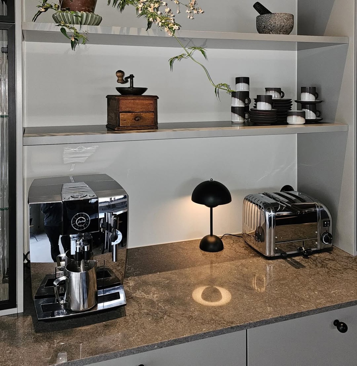 Coffee machine, toaster, black lamp, vintage grinder, cups, plates, and potted plants on shelves.