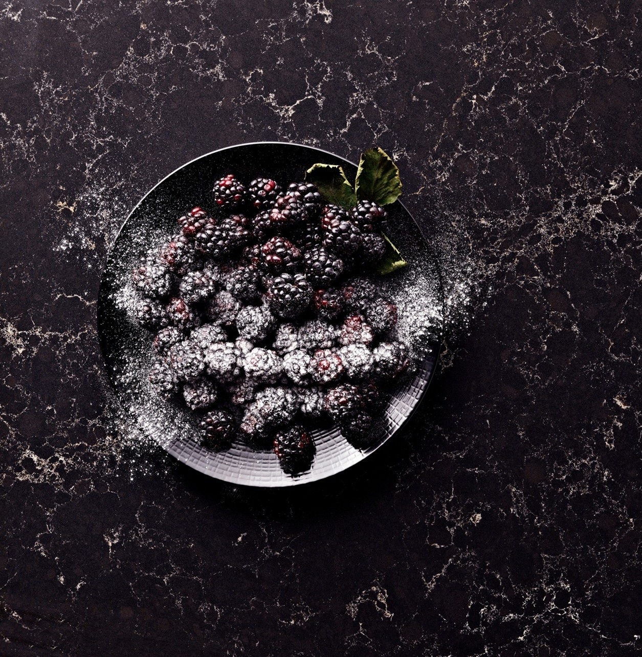 Blackberries dusted with powdered sugar on a dark plate, garnished with a leaf.