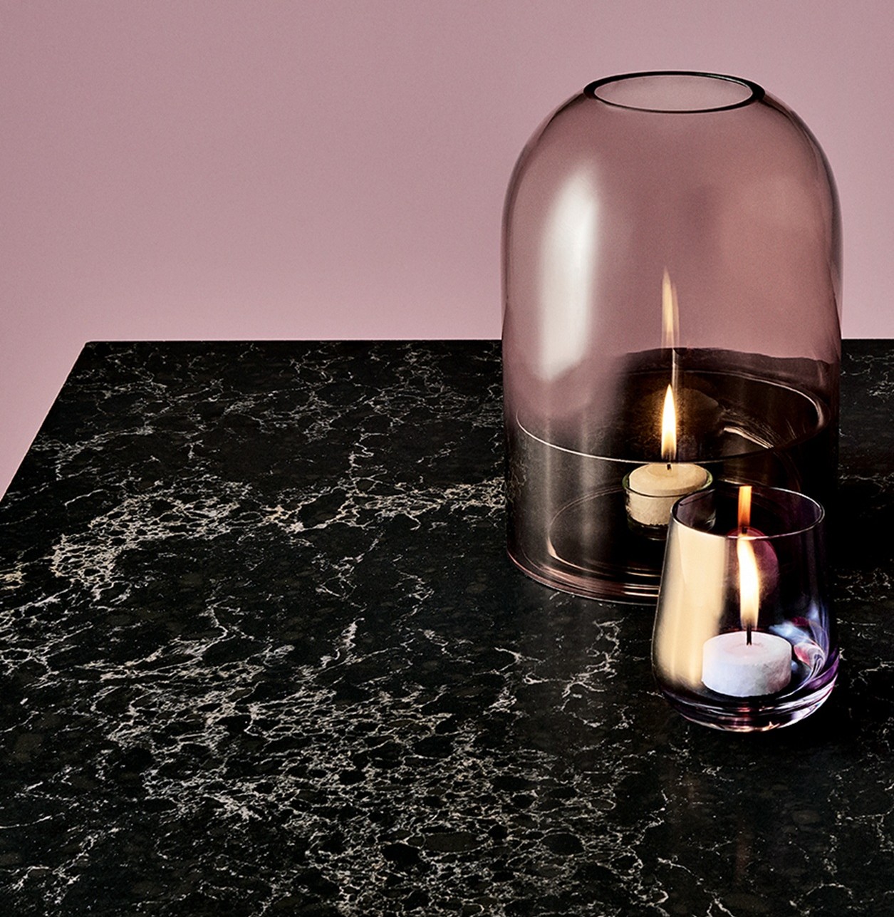 Two candles in glass holders on black marble surface with a glass dome